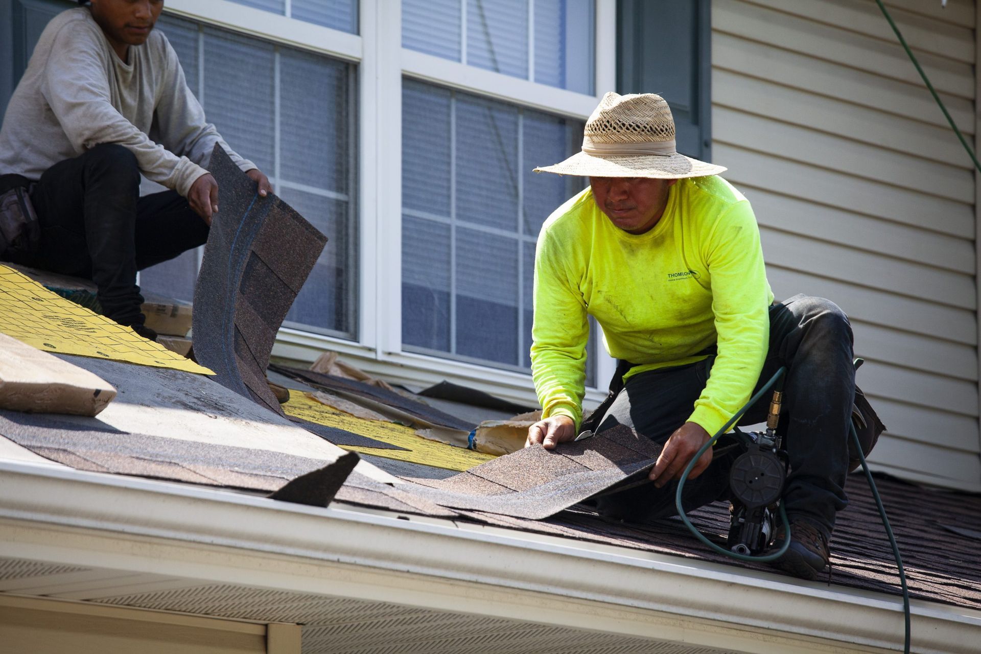 Two construction workers wearing safety gear install asphalt roof shingles on a residential home.