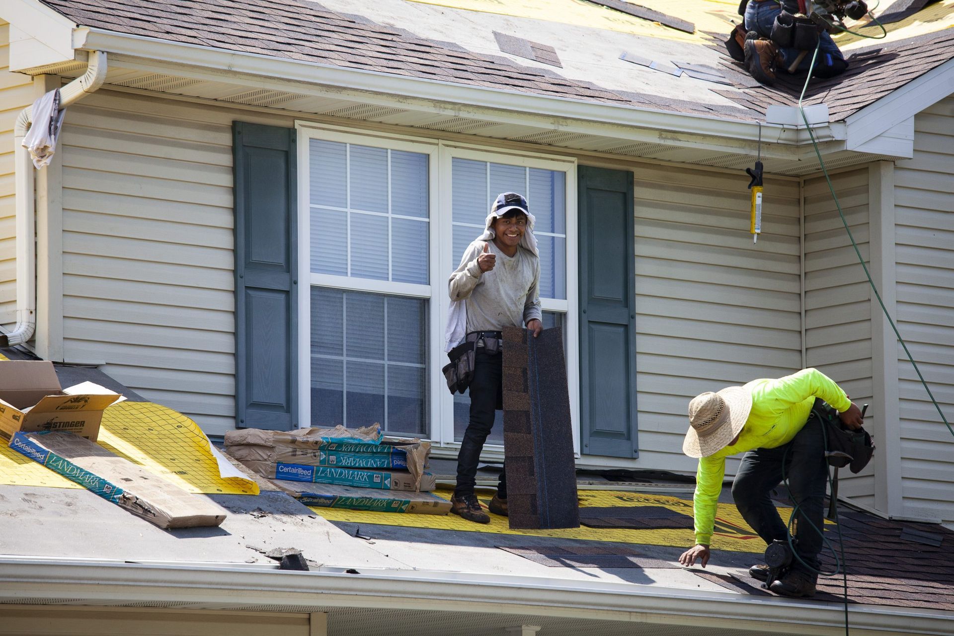 Two workers install shingles on a residential roof, with one standing and one crouching to work.