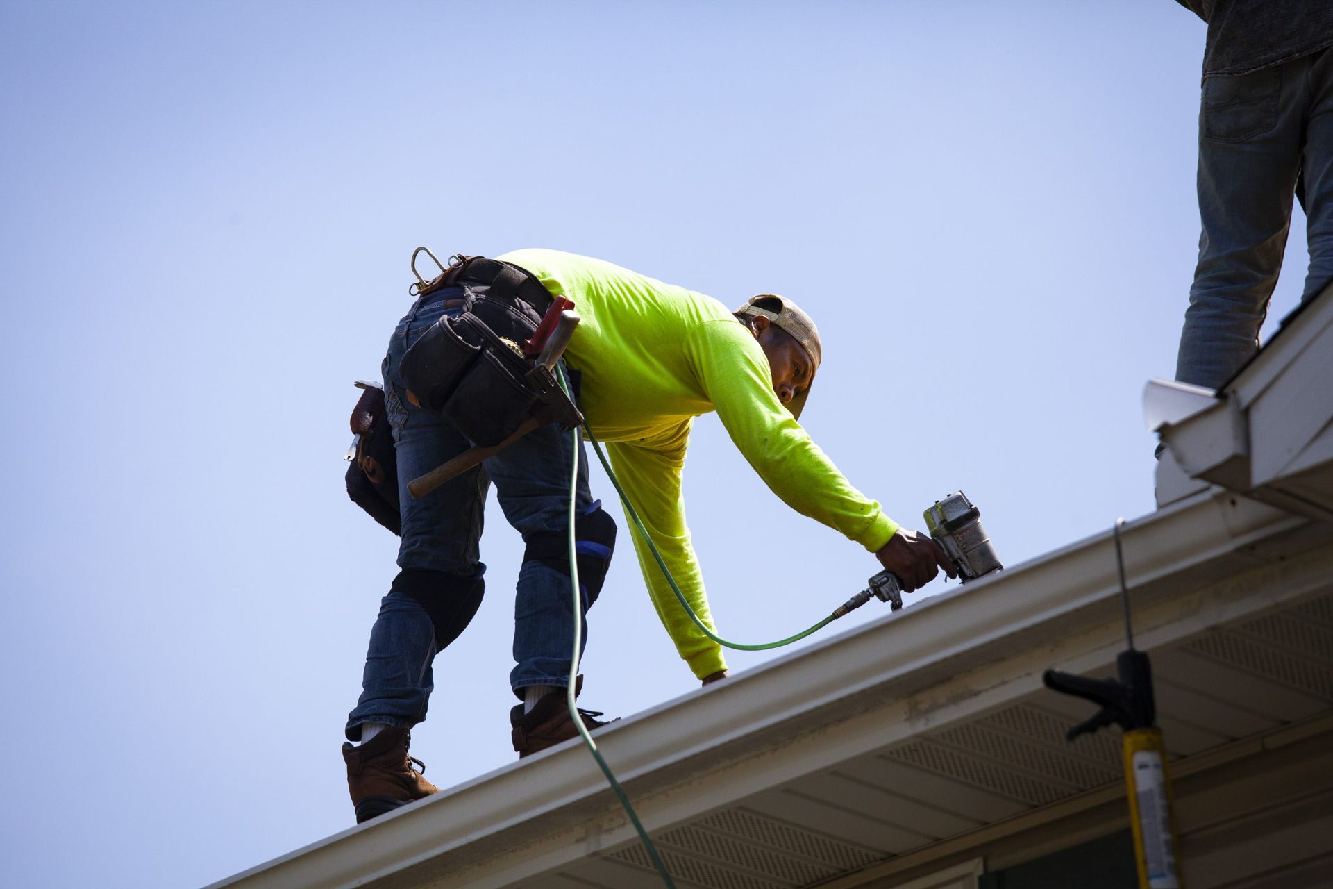 A construction worker in a neon yellow shirt uses a nail gun on a roof edge against a clear blue sky.