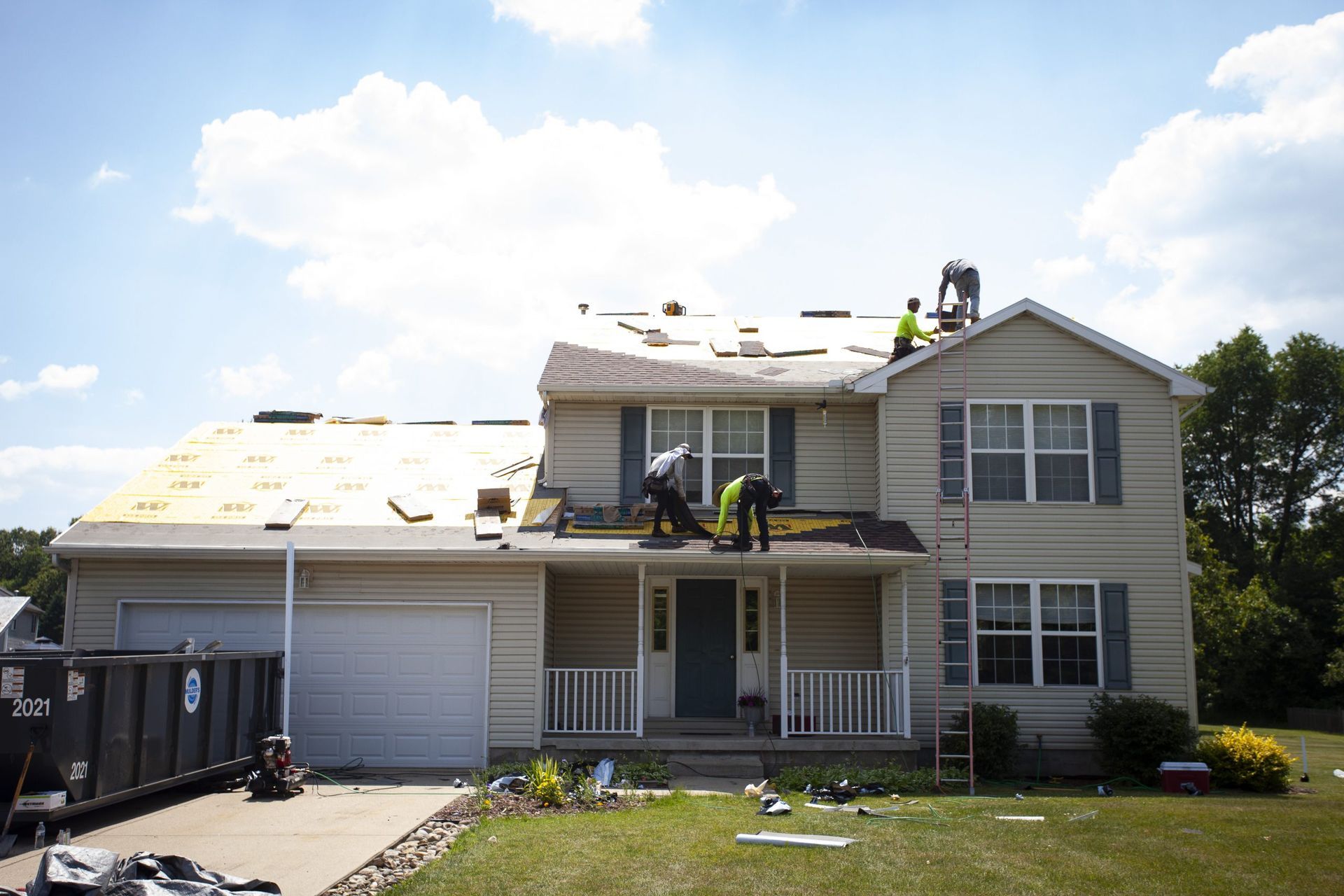 Construction workers replacing the roof of a two-story beige house with a dumpster parked in the driveway.