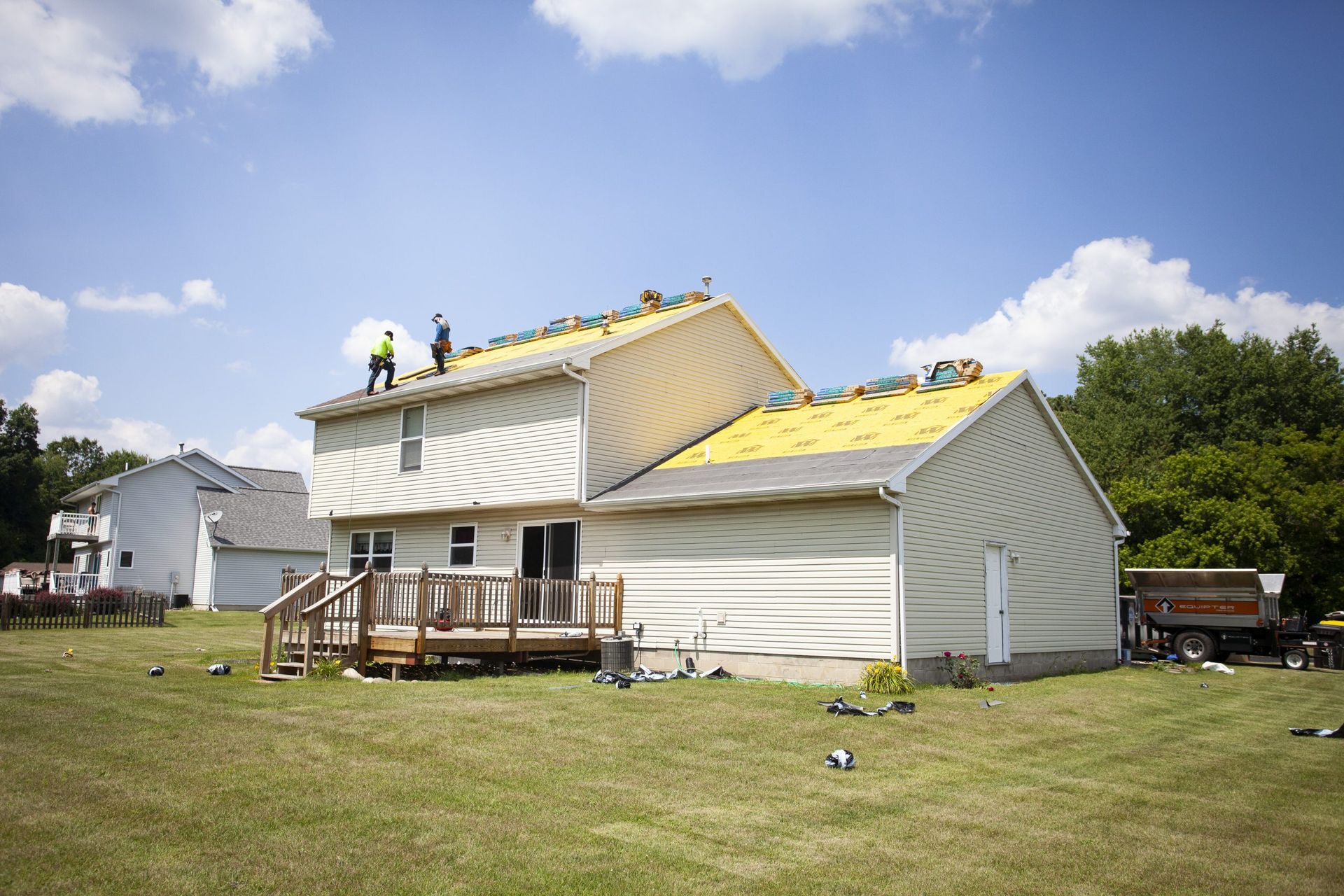 Roofers work on the yellow-sheathed roof of a two-story beige house on a sunny day with a green lawn and blue sky.