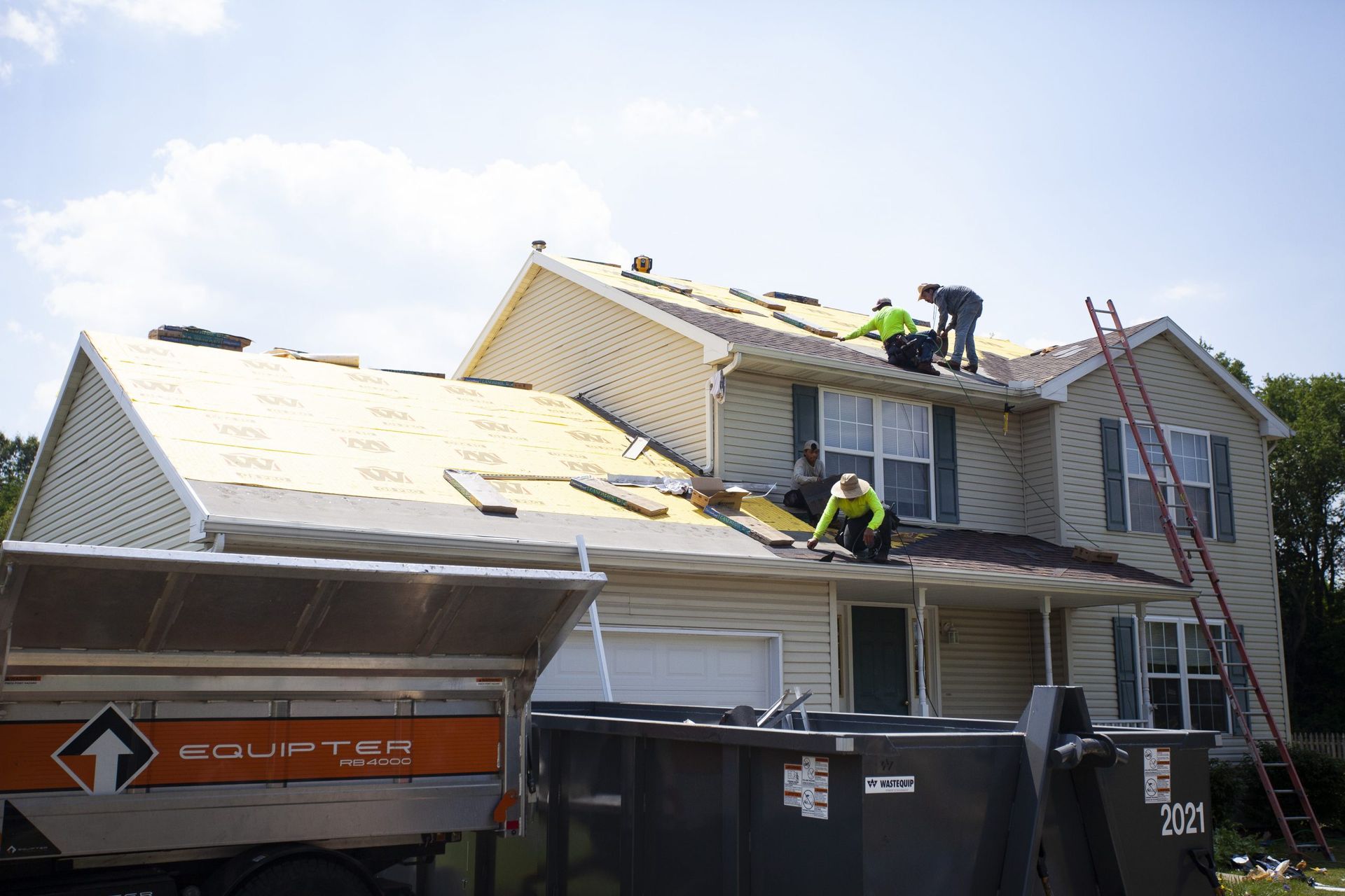 Roofers working on the shingles and decking of a tan two-story house with a dumpster and equipment trailer in the yard.