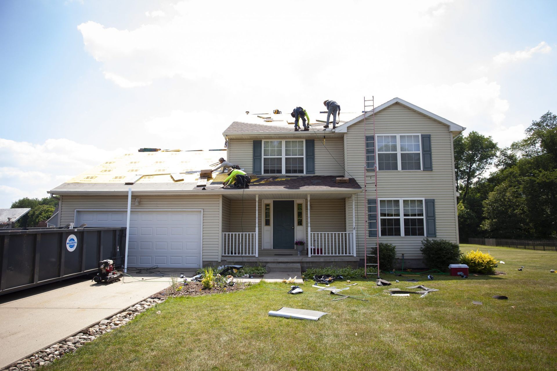 Workers on the roof of a two-story beige house undergoing repairs, with a dumpster in the driveway.
