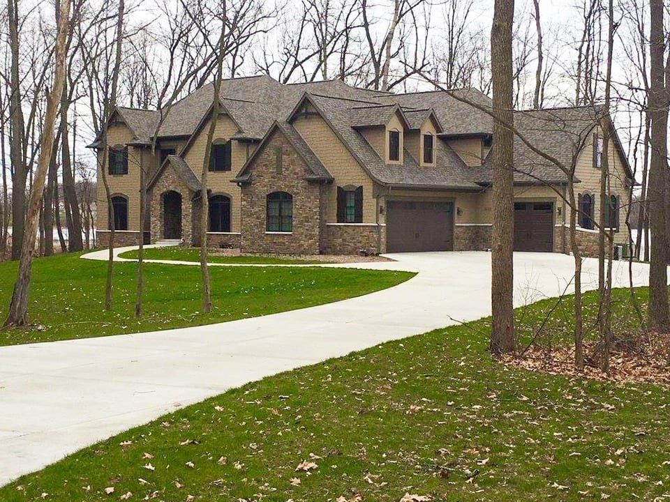 A two-story tan and stone-veneer house with a dark shingled roof, a paved driveway, and surrounding bare trees.