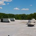 Two rectangular HVAC units and two circular roof vents sit on a flat gray roof under a blue, cloudy sky.