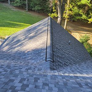 High-angle view of a grey shingled roof with a central ridge vent, surrounded by trees and green lawn.