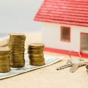 Stacks of coins and keys placed in front of a small model house on a beige surface.