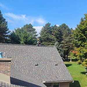 A person working on the shingled roof of a house on a sunny day with trees in the background.