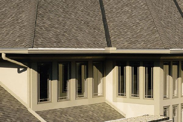 A modern, tan-colored home exterior with an asphalt shingled roof, overhanging eaves, and vertical windows.