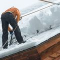 A person in an orange jacket and black pants uses a long tool to clear deep snow from a wooden roof.