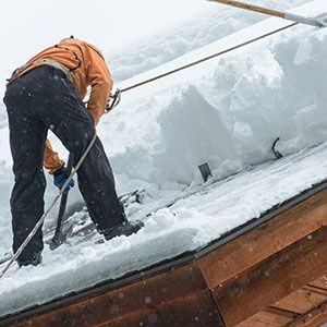 A person in an orange jacket and black pants uses a long tool to clear deep snow from a wooden roof.