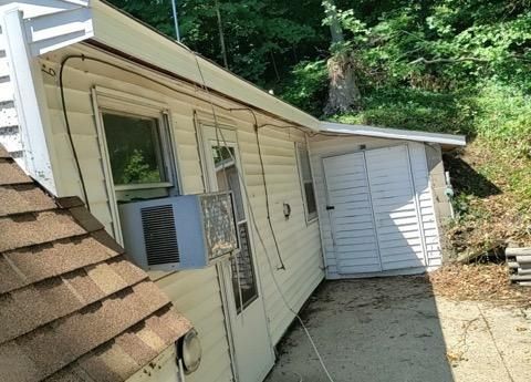A light-colored house exterior with a window air conditioner unit and a white door, set against a wooded hillside.