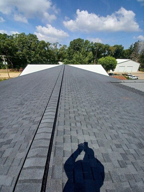 A rooftop view of dark gray asphalt shingles along a ridge, with a white roof section and trees in the distance.