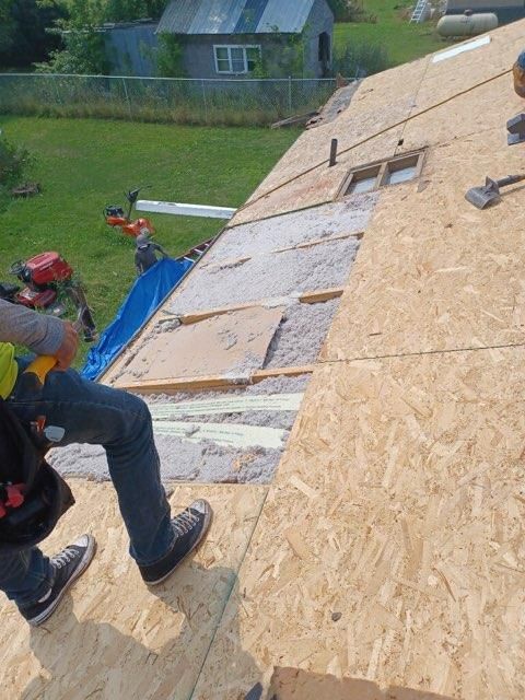 A worker stands on a partially sheathed roof exposing insulation and framing, with a yard visible in the background.