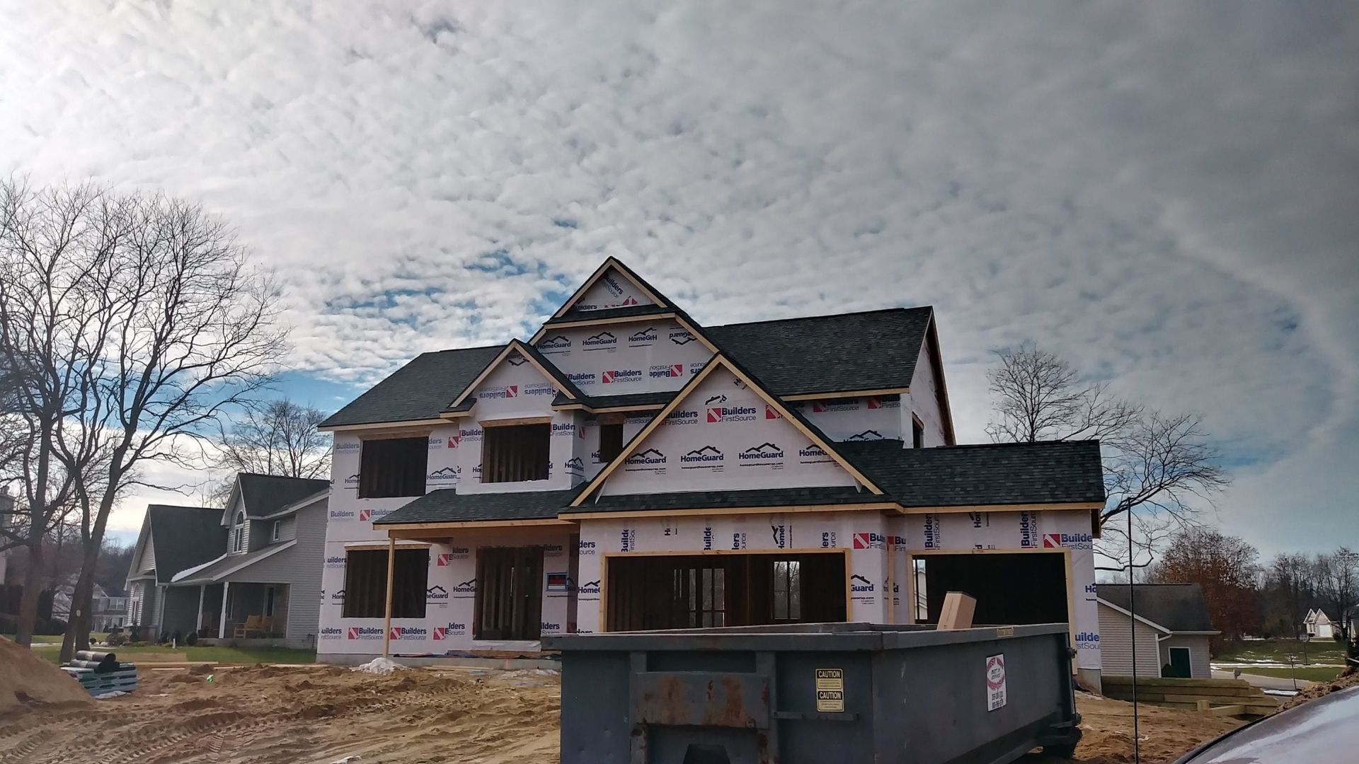 A two-story house under construction with a protective wrap, shingled roof, and a large dumpster in the foreground.