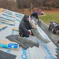 Four people in hoodies install gray shingles onto a house roof covered in branded underlayment paper outdoors.