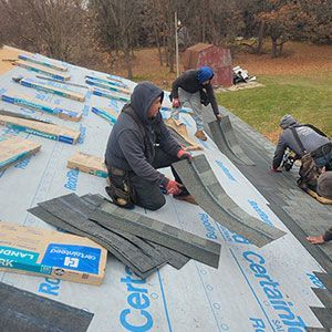 Four people in hoodies install gray shingles onto a house roof covered in branded underlayment paper outdoors.