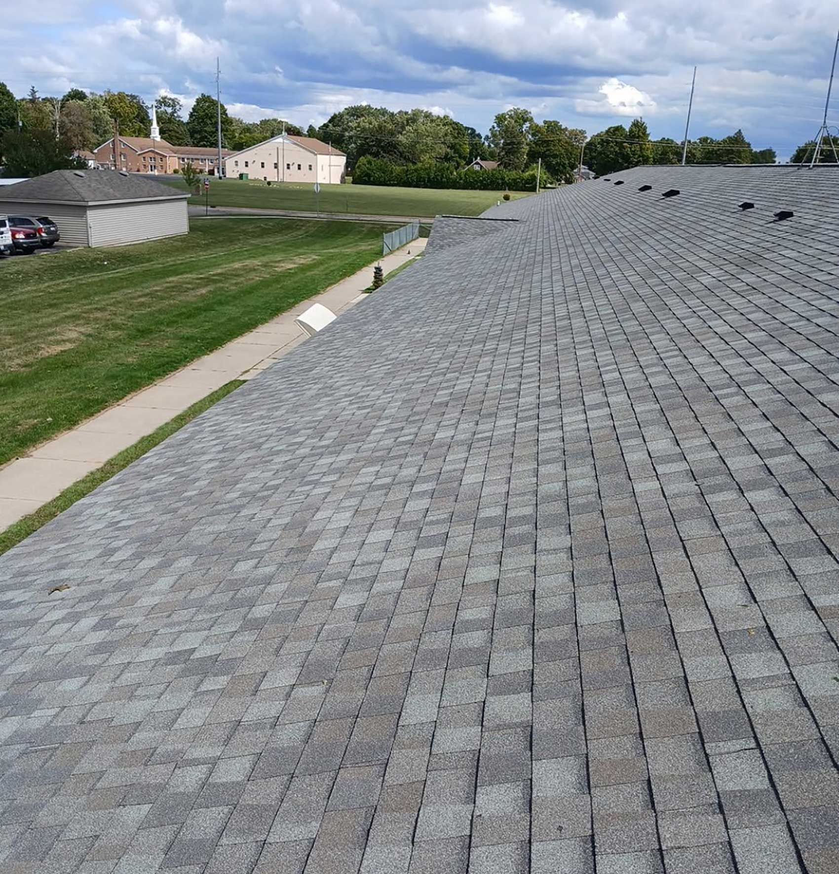 A high-angle view of a gray asphalt shingle roof, with a grassy lawn, sidewalk, and buildings visible in the distance.