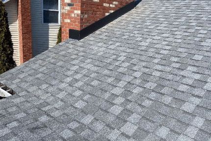 A sloped residential roof covered in gray asphalt shingles, with a brick chimney and siding in the background.