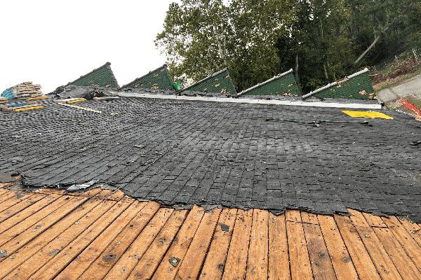 A partially stripped roof shows wooden slats in the foreground and a layer of black roofing felt against green peaks.