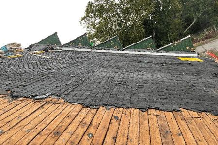 A partially stripped roof shows wooden slats in the foreground and a layer of black roofing felt against green peaks.