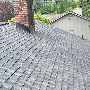 A close-up view of a shingled roof with a brick chimney and a light-colored house in the background.