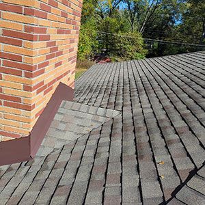 A brick chimney rises from a shingled roof, featuring brown metal flashing installed at the base to prevent water leaks.