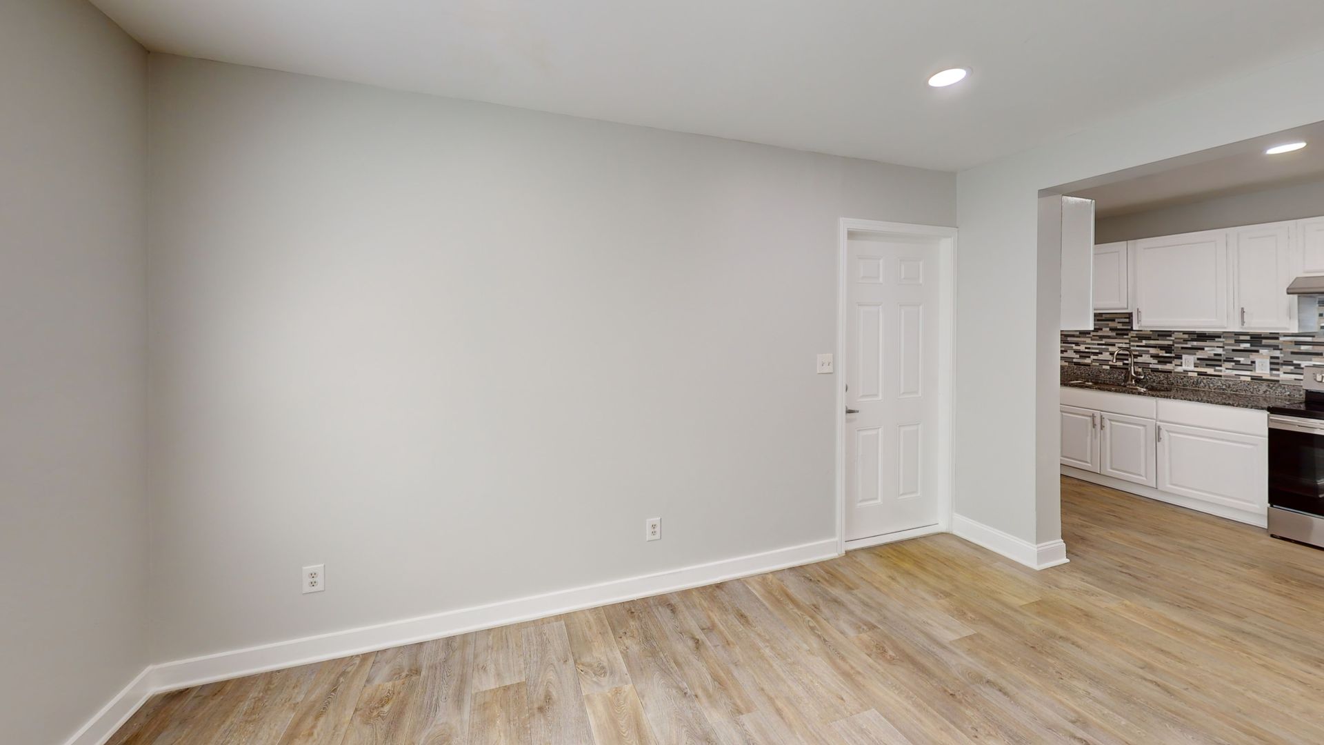 Empty modern room with gray walls, light wood floors, and a view into a kitchen area