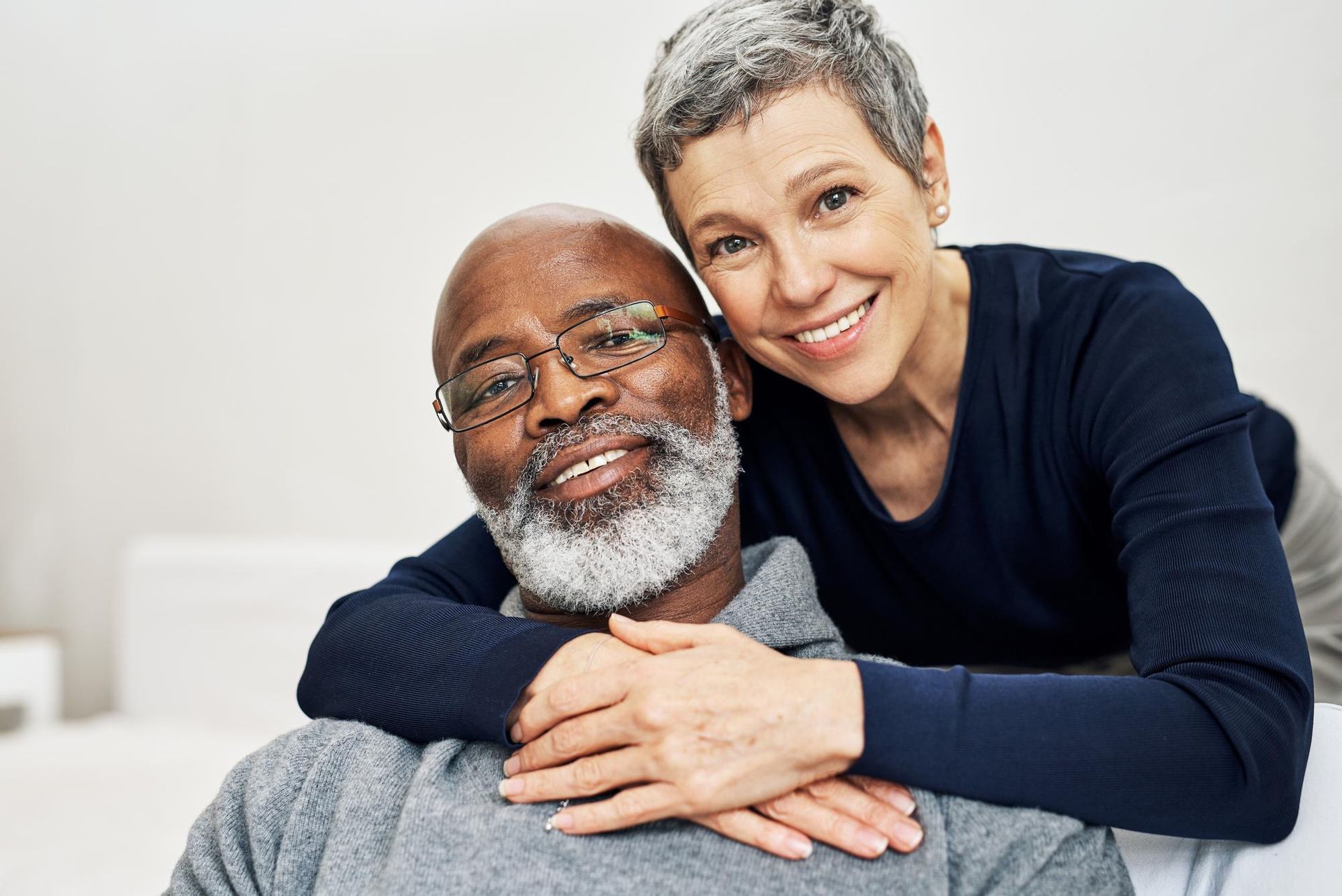 Elderly couple smiles, embracing. Woman's arms around man's shoulders. White and gray hair.