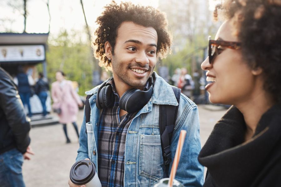 Smiling Black man and woman converse outdoors. Man holds coffee, woman wears sunglasses.
