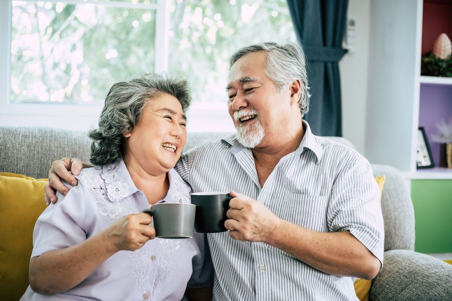 Smiling senior couple, embracing on a sofa, holding mugs of coffee indoors.