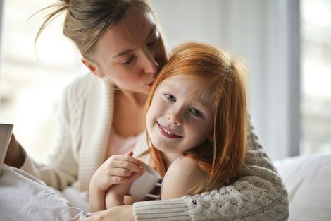 Woman kissing a smiling red-haired girl. They are indoors, likely in a bed, looking happy.