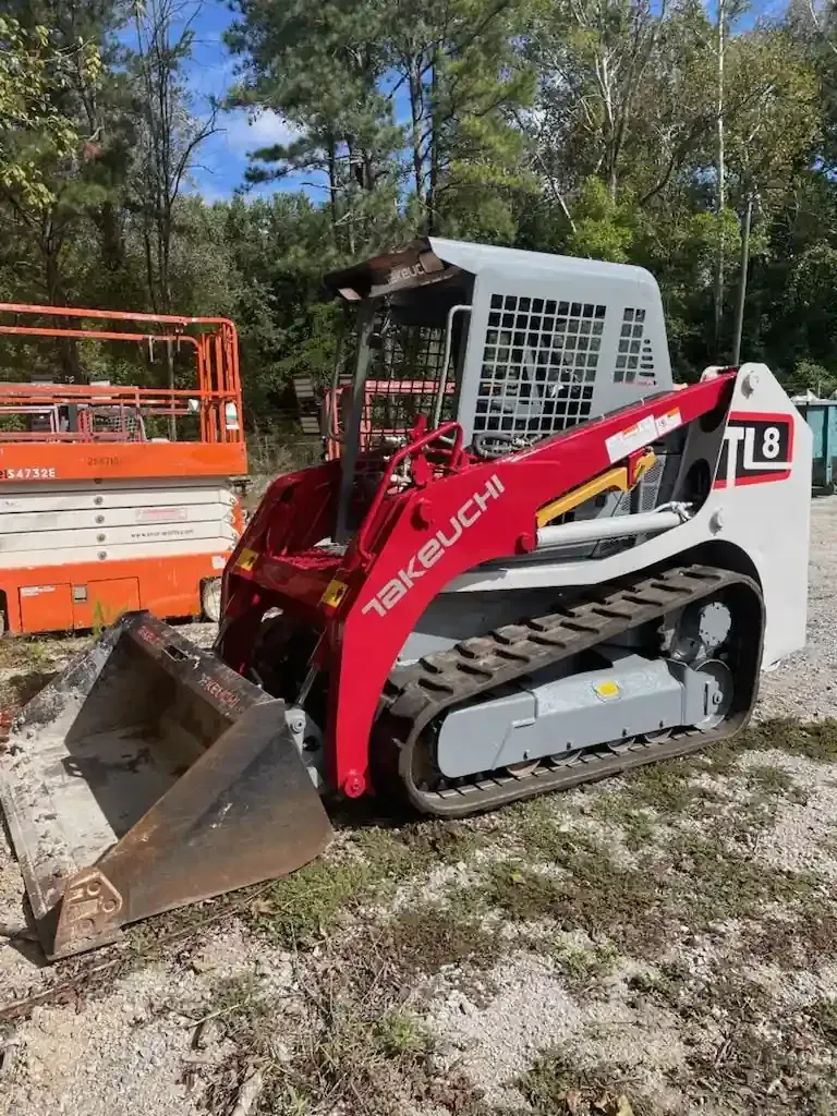 Red Takeuchi compact track loader with front bucket on a dirt site, trees in the background.