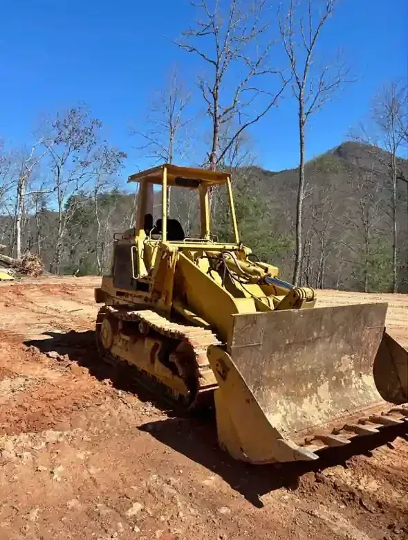 Yellow bulldozer on a dirt road clearing land, with bare trees and hills in the background.