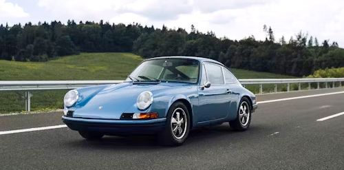 Blue Porsche 911 driving on a highway, with green trees in the background and a cloudy sky.