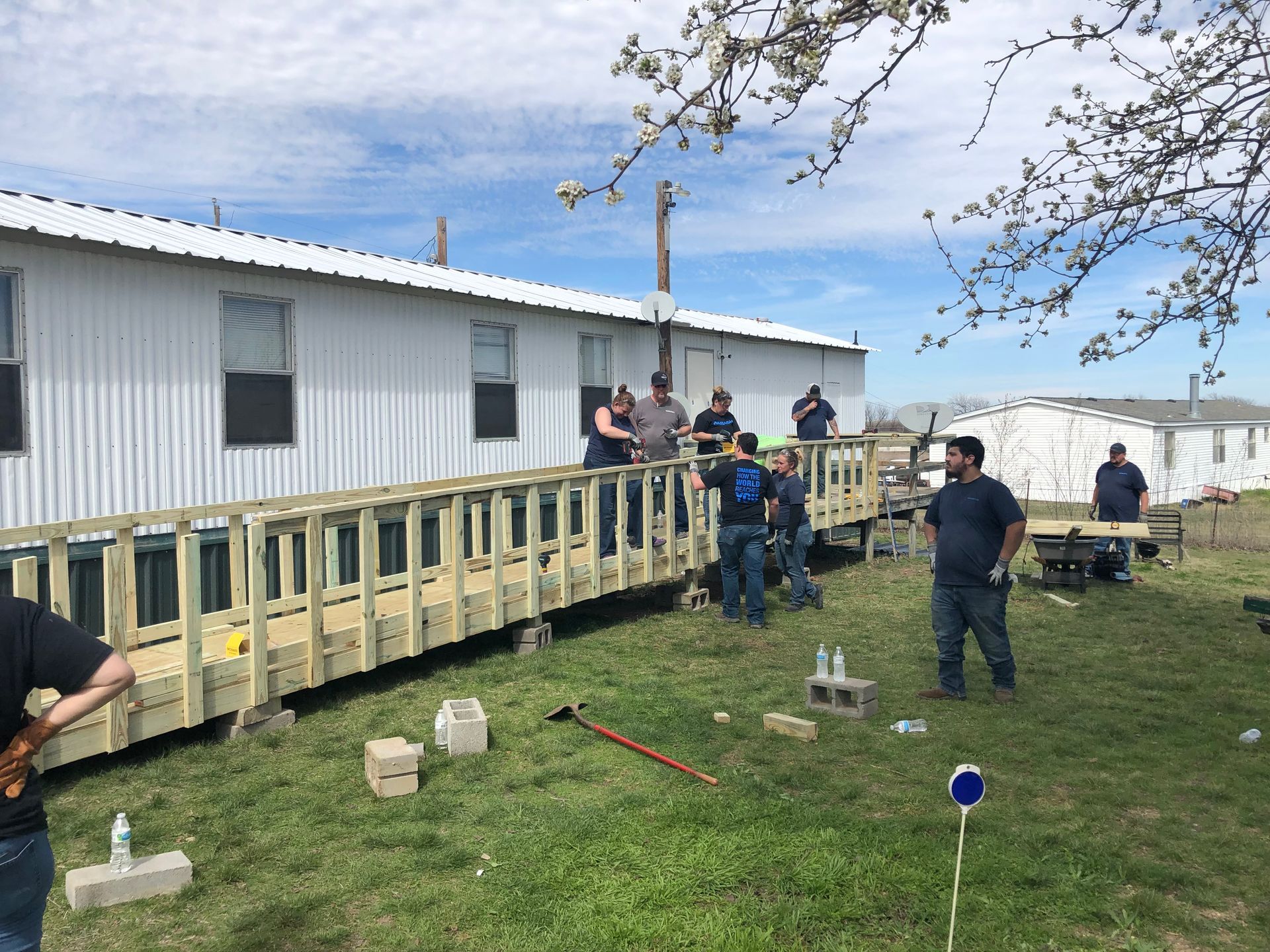 People standing on a wooden ramp outside a white building on a grassy lot, with tools scattered nearby