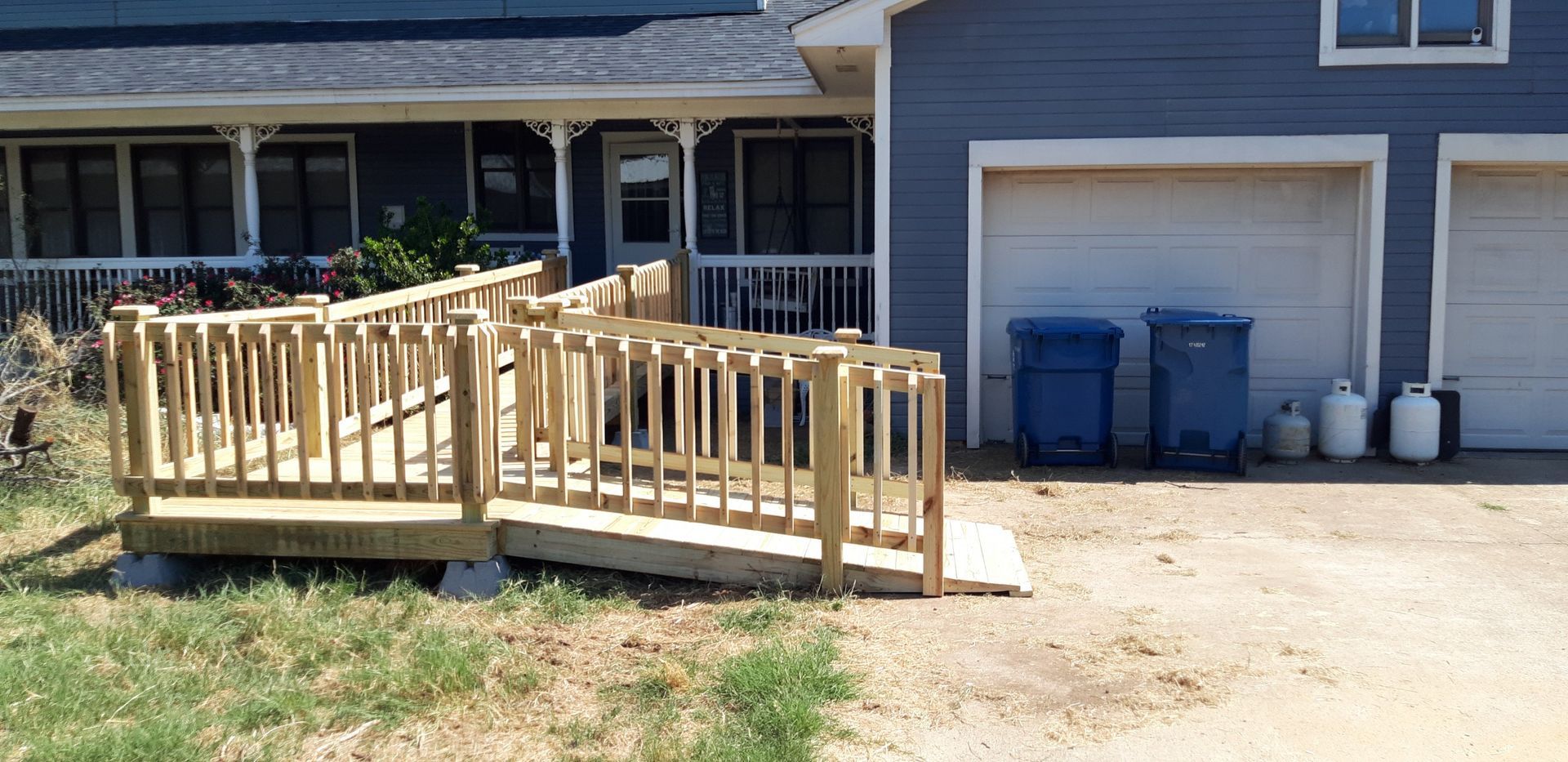 Wooden accessibility ramp leading to a house entrance beside two blue trash bins and a garage
