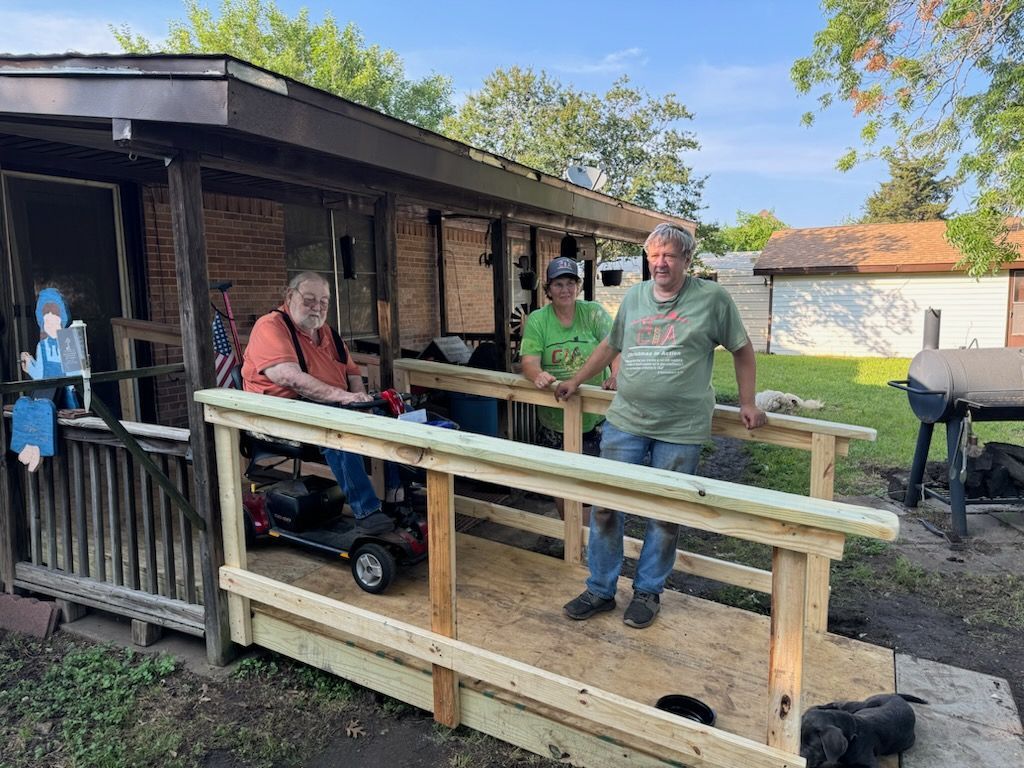 People on a wooden ramp outside a small cabin, with one man leaning on the railing and a wheelchair nearby