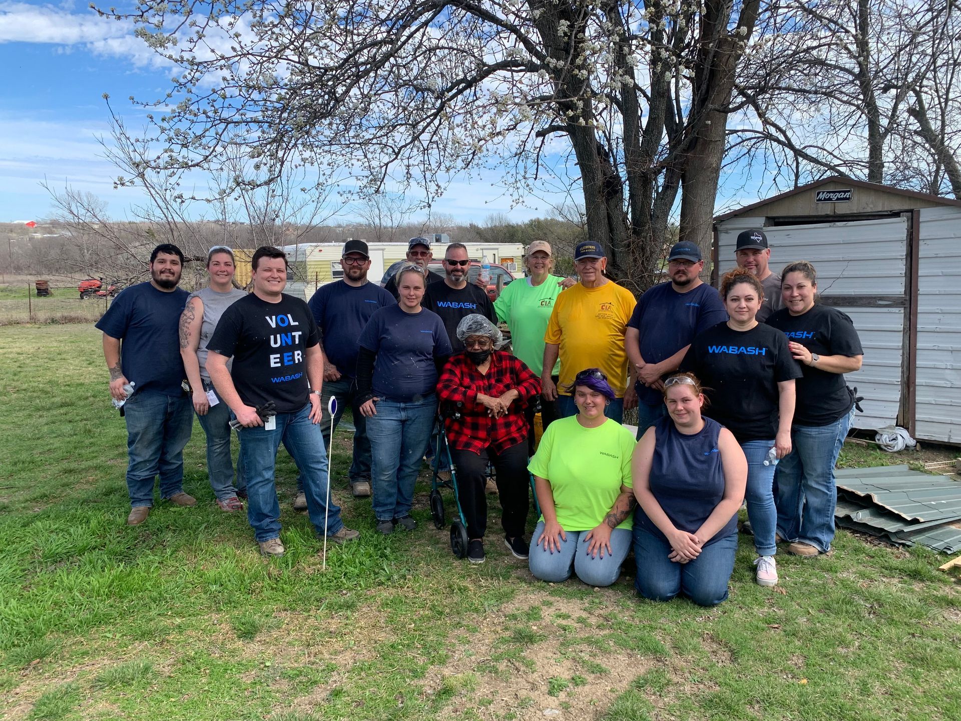 Group of people posing outdoors by a tree and shed, with one person kneeling in bright green and others standing.