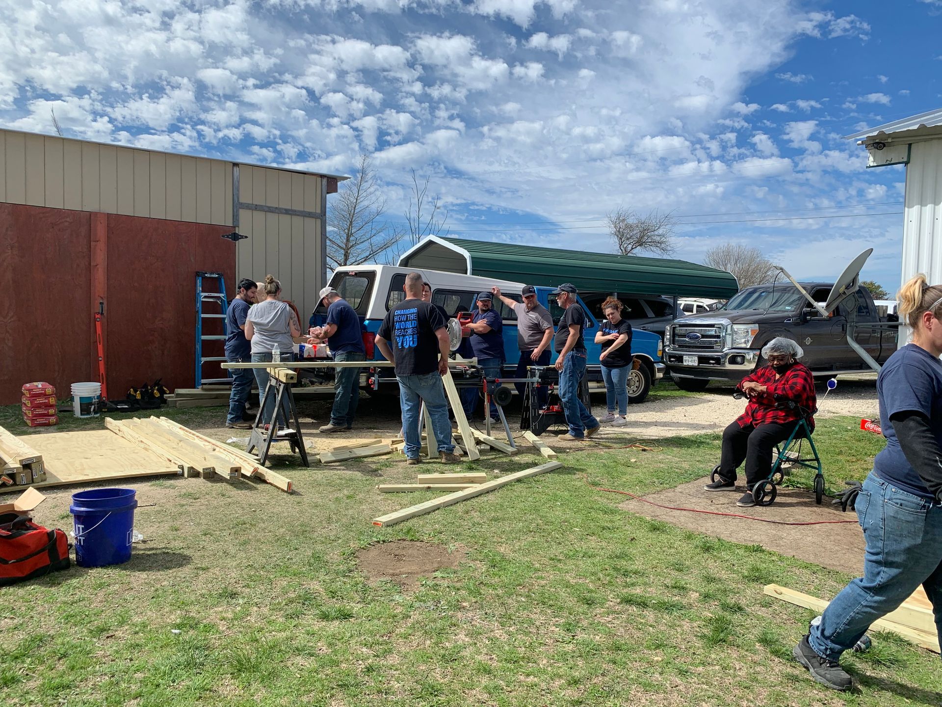 People gathered outdoors beside a barn, working around a canopy and table under a blue sky.