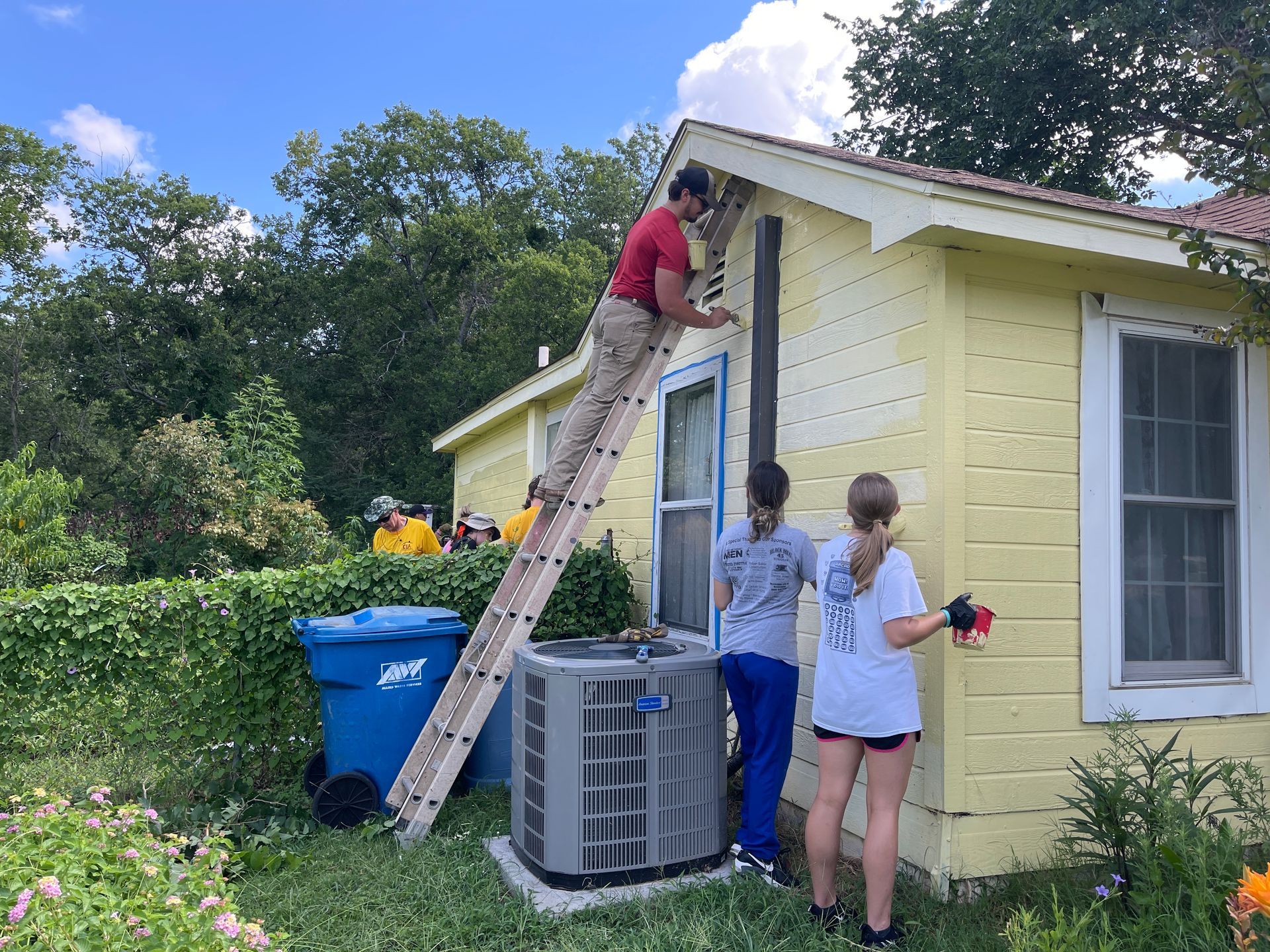 Two people repairing a yellow house roof, one on a ladder and one standing below with tools.