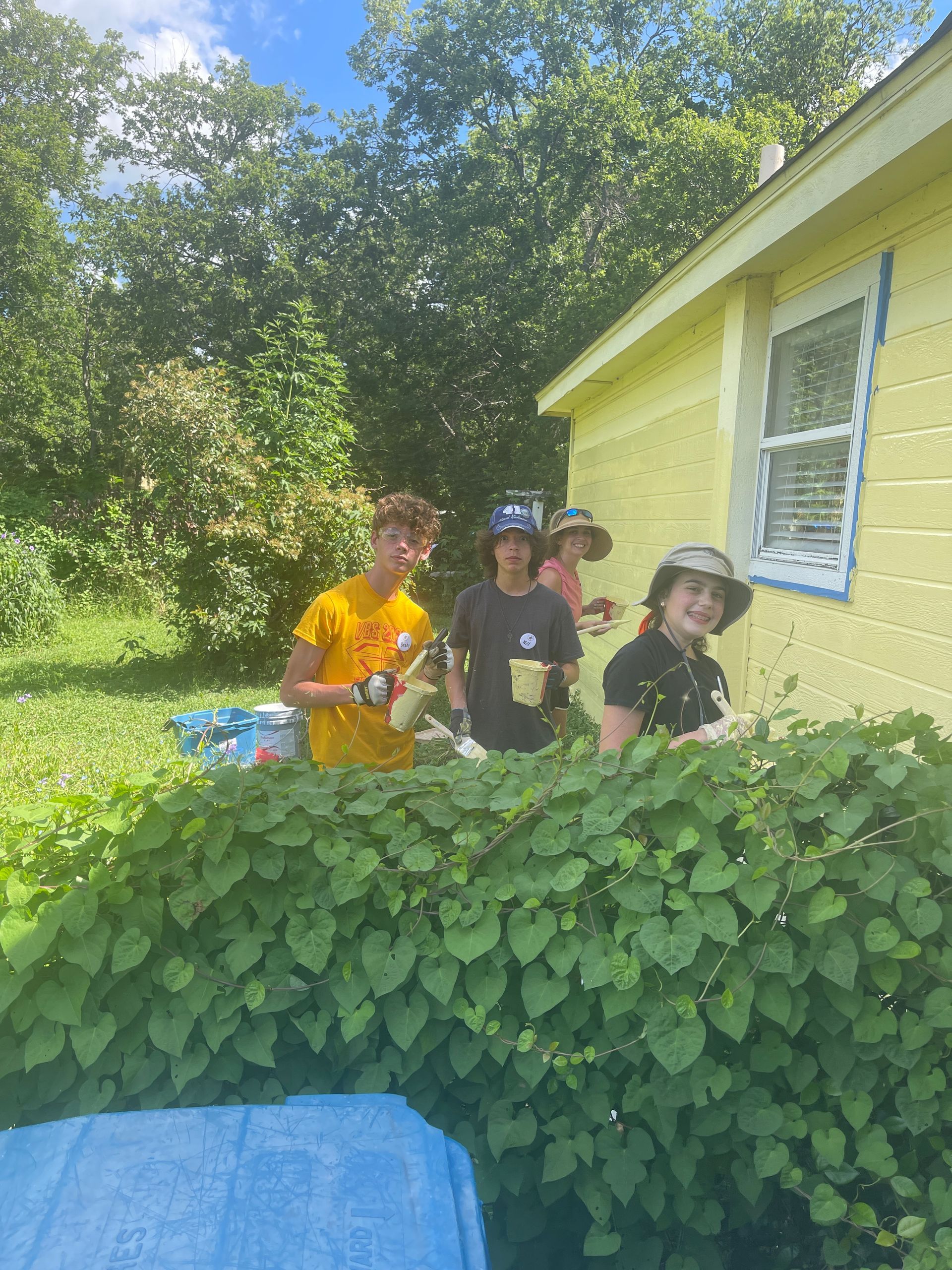 Three people standing behind a leafy hedge beside a yellow house on a sunny day