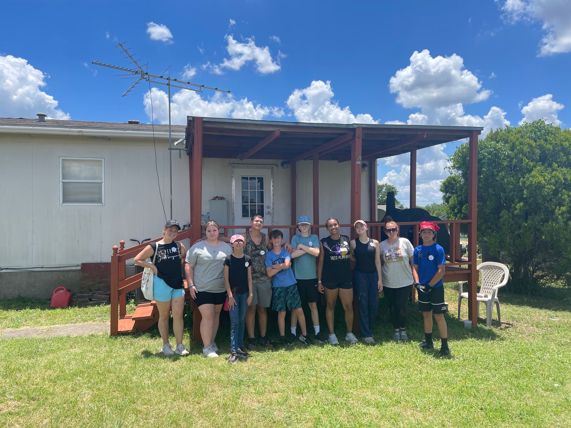 Group of people standing on a grassy lawn in front of a white building and wooden porch under a blue sky.