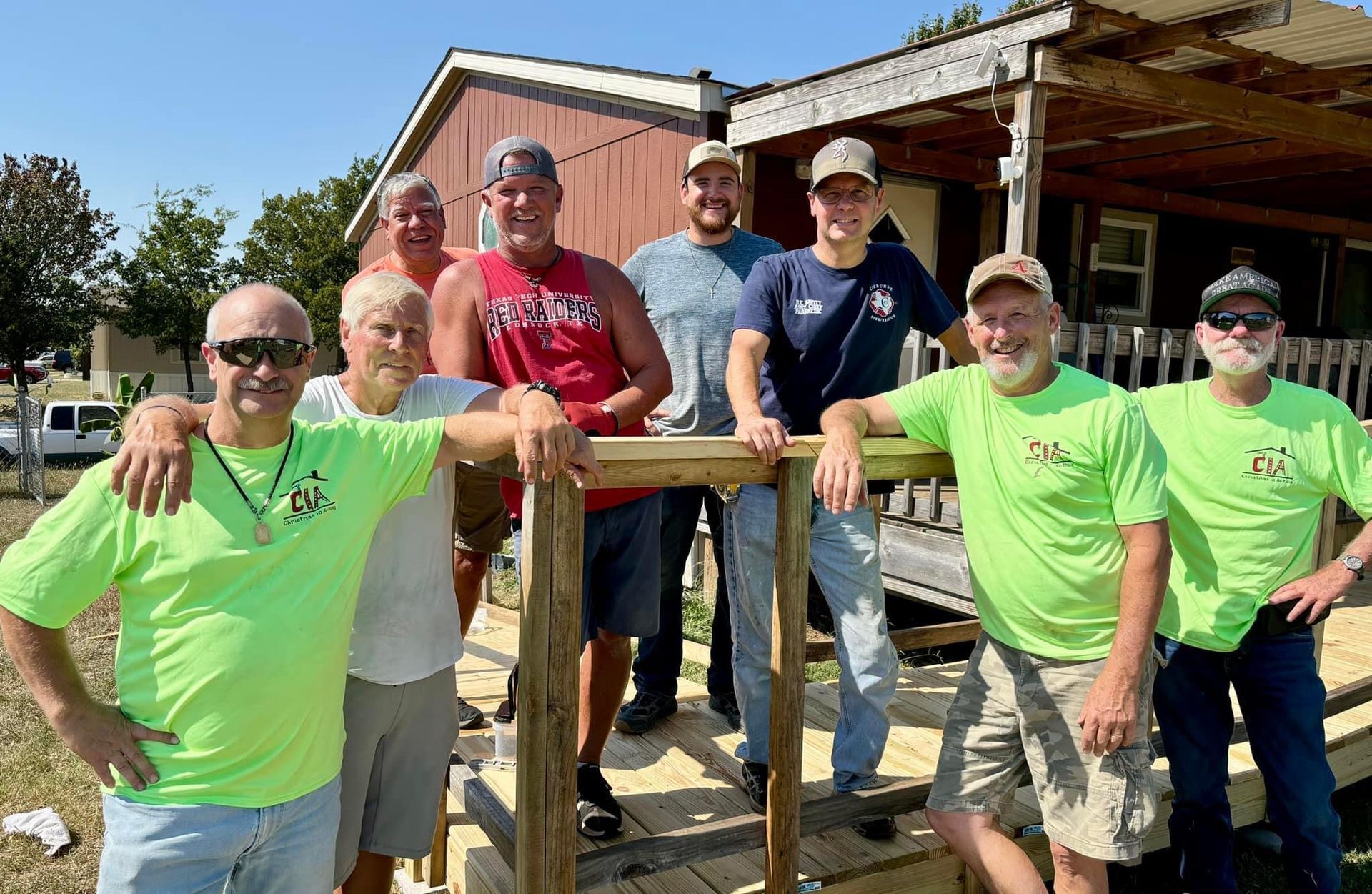 Group of volunteers posing outdoors beside a wooden porch, smiling in bright neon green shirts.