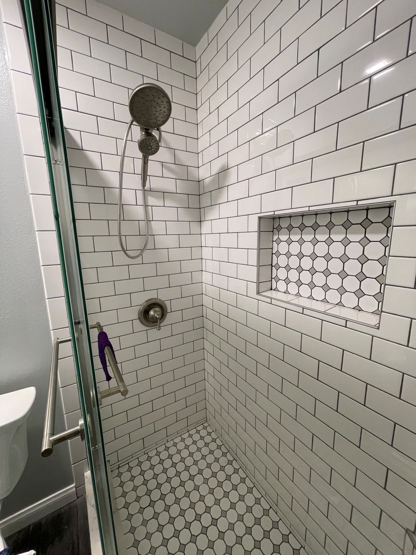 A shower with white subway tile walls, a mosaic tile floor and recessed shelf, and a glass door with a silver handle.