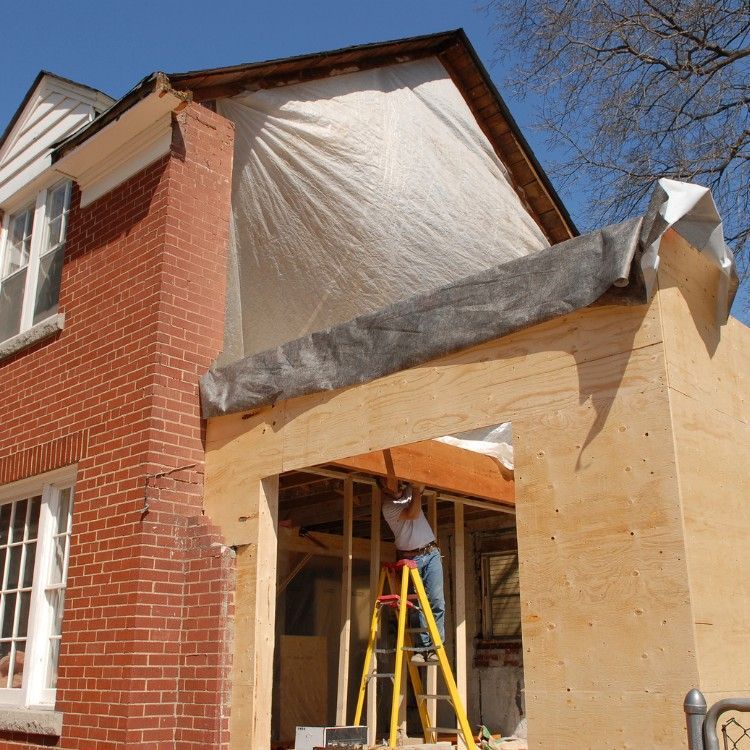A worker stands on a ladder under a plastic-covered wood frame addition attached to a brick house under construction.