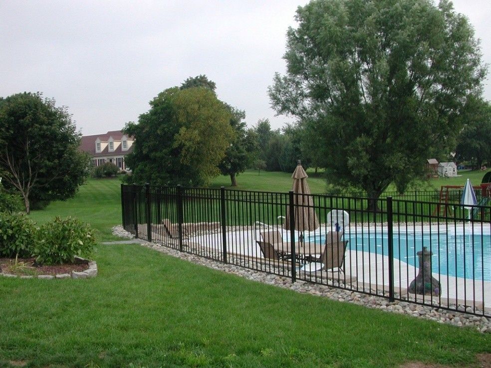 Black fence surrounds a swimming pool in a green yard with trees and a house in the background.