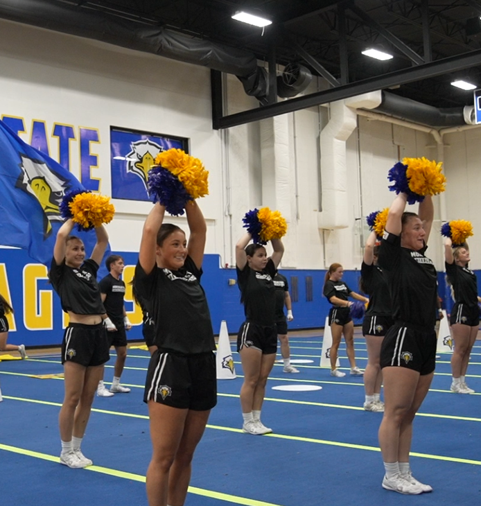 Cheerleaders in formation holding Blue and Gold poms with a Morehead Flag in the background.