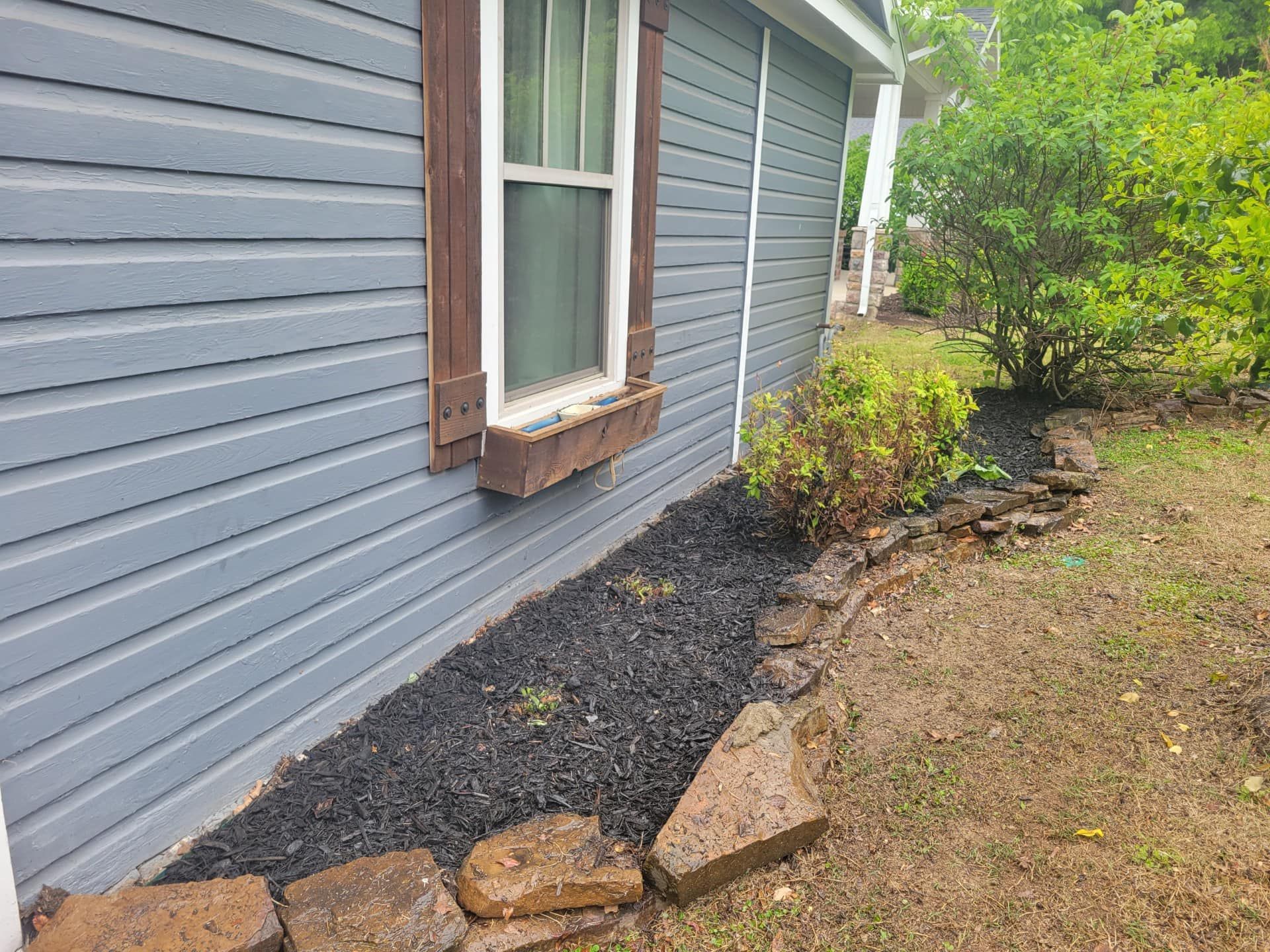 Blue house with a brown-trimmed window and a dark mulch flower bed lined with brown stones.