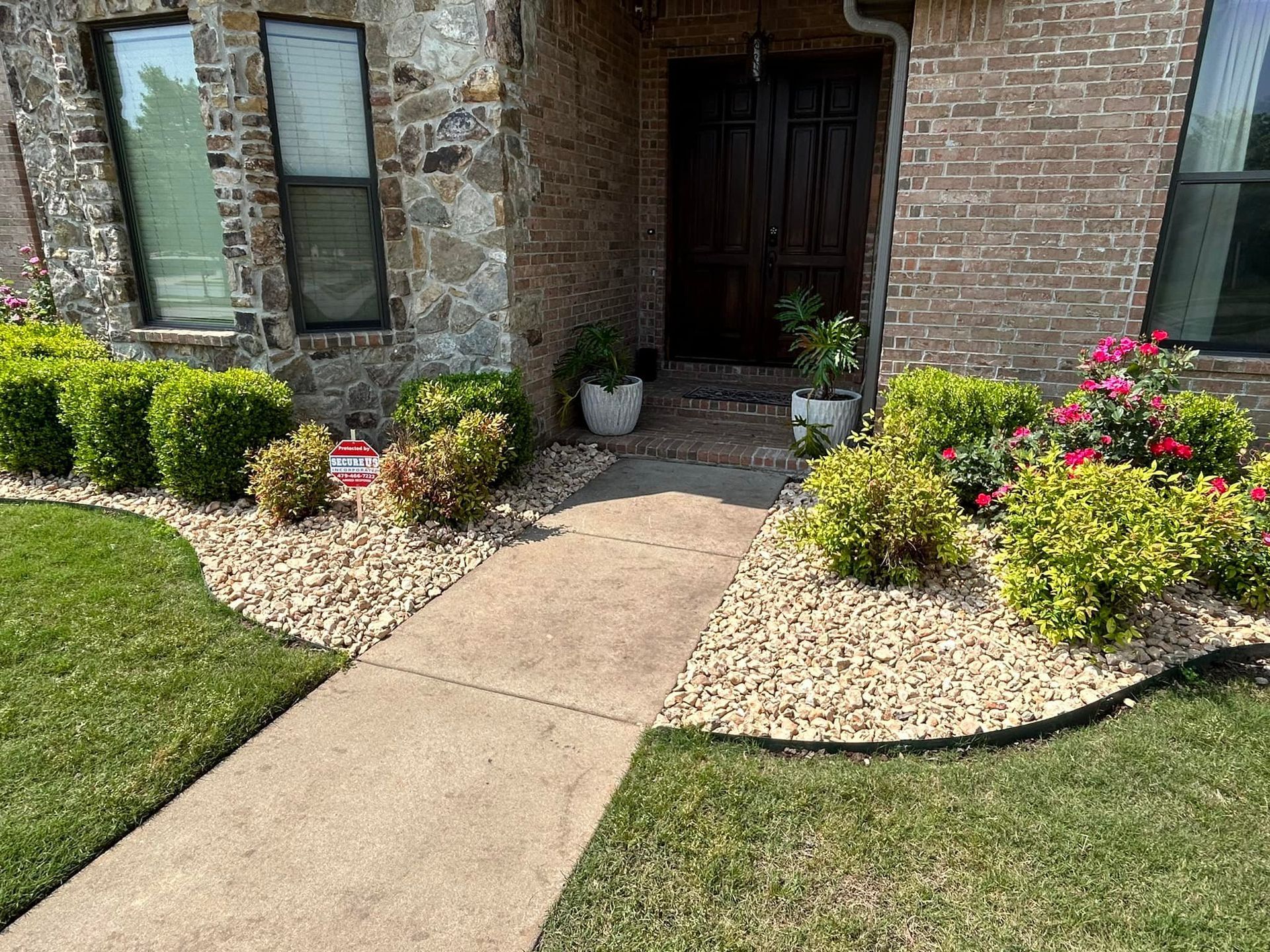 Concrete walkway leading to a house with stone facade, brick exterior, and landscaped flower beds with green plants and red flowers.